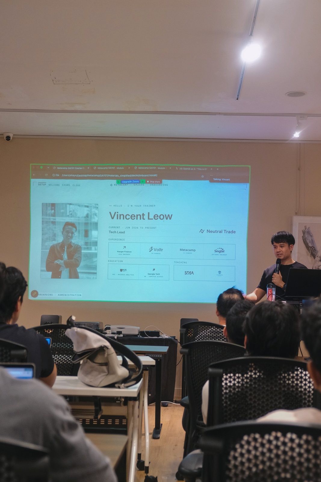 A trainer speaks to the class with a microphone beside a projected slide of his profile, learners watching from their seats
