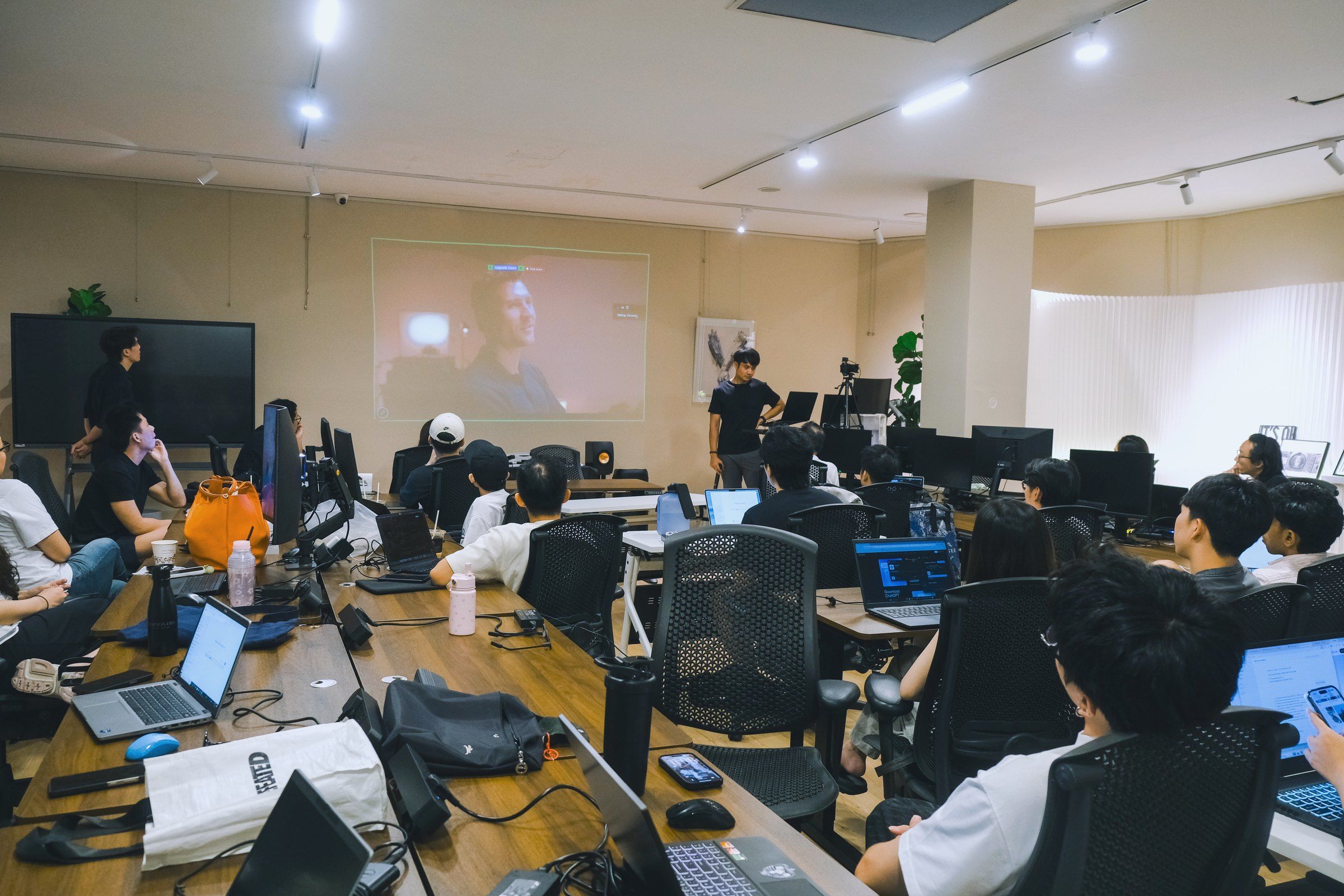 A full classroom of learners at their laptops, facing a projected video at the front of the room