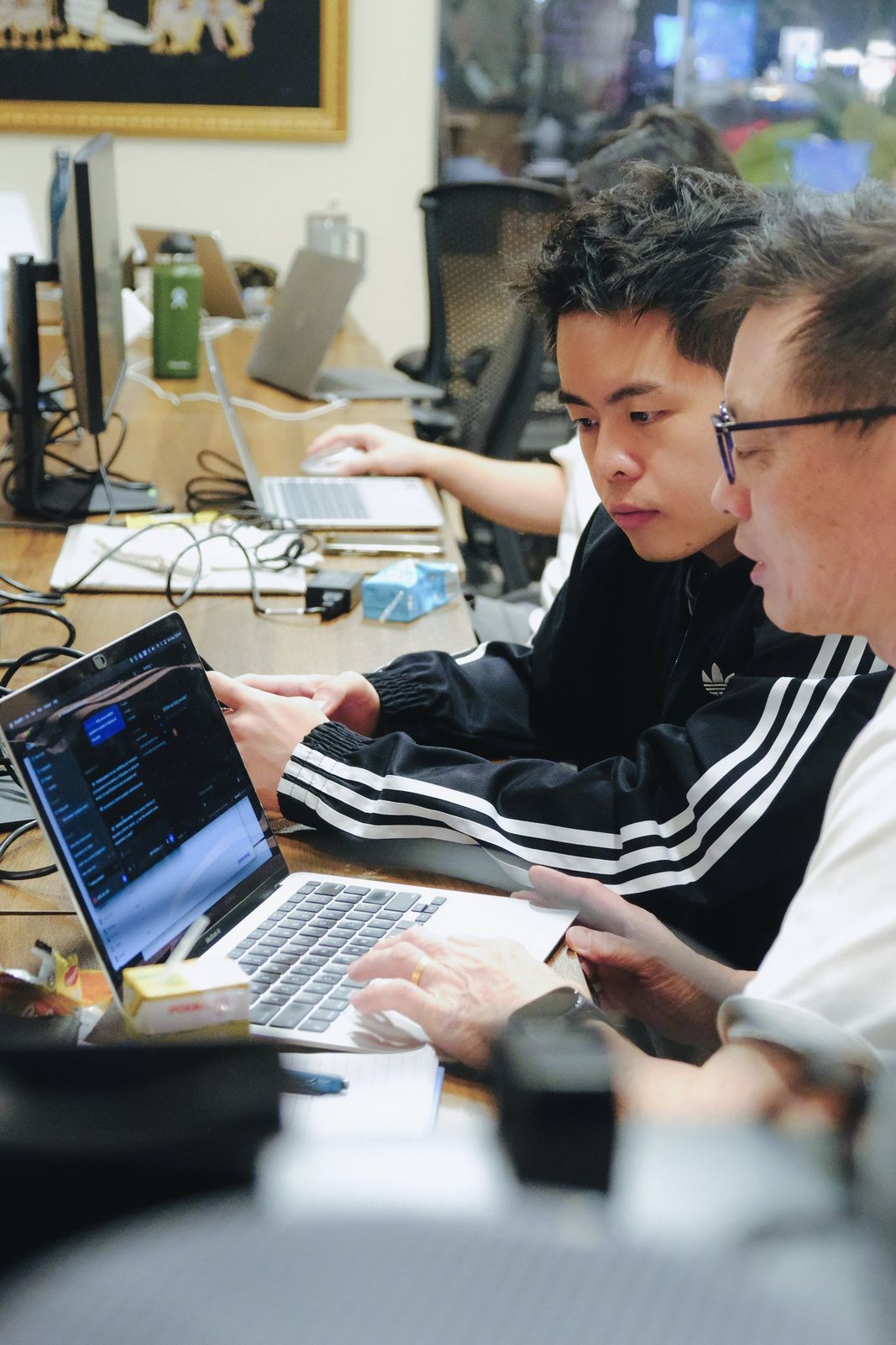 Two learners lean in together over one laptop at a long class table