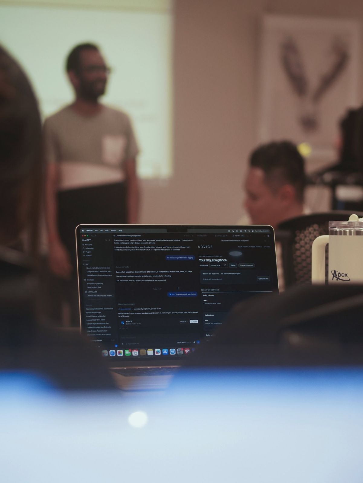 A laptop open to an AI tool while the instructor speaks at the front of the room